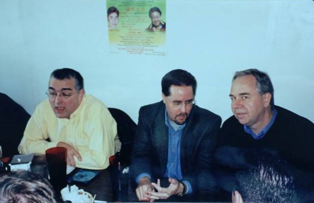 An old print of three men seated side by side at a Pho brunch table against a white wall, one in a yellow shirt talking, one in a dark jacket and glasses, one in a dark sweater, with a poster on the wall above them.