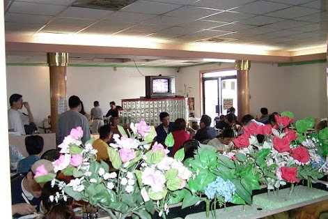 The dining room at Pho 87 during a Pho brunch, with pink and red silk flowers in the foreground, a row of tables full of diners, gold pillars and a small television on the far wall.