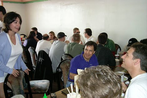 A Sunday brunch at Pho 87: a woman in a light blue cardigan stands at the end of a crowded table while friends sit talking over bowls of pho, with more tables of diners behind them.